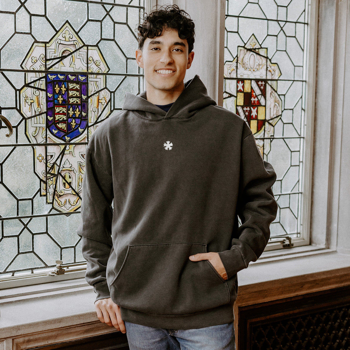 College-aged man wearing the Sigma Nu embroidered black hoodie, standing in front of stained glass windows with fraternity-style crest designs.