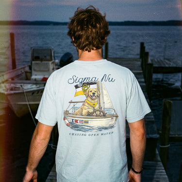 Young man standing on a dock at sunset wearing a light blue t-shirt featuring a golden retriever steering a sailboat with a flag and the phrase “Chasing Open Water” on the back.