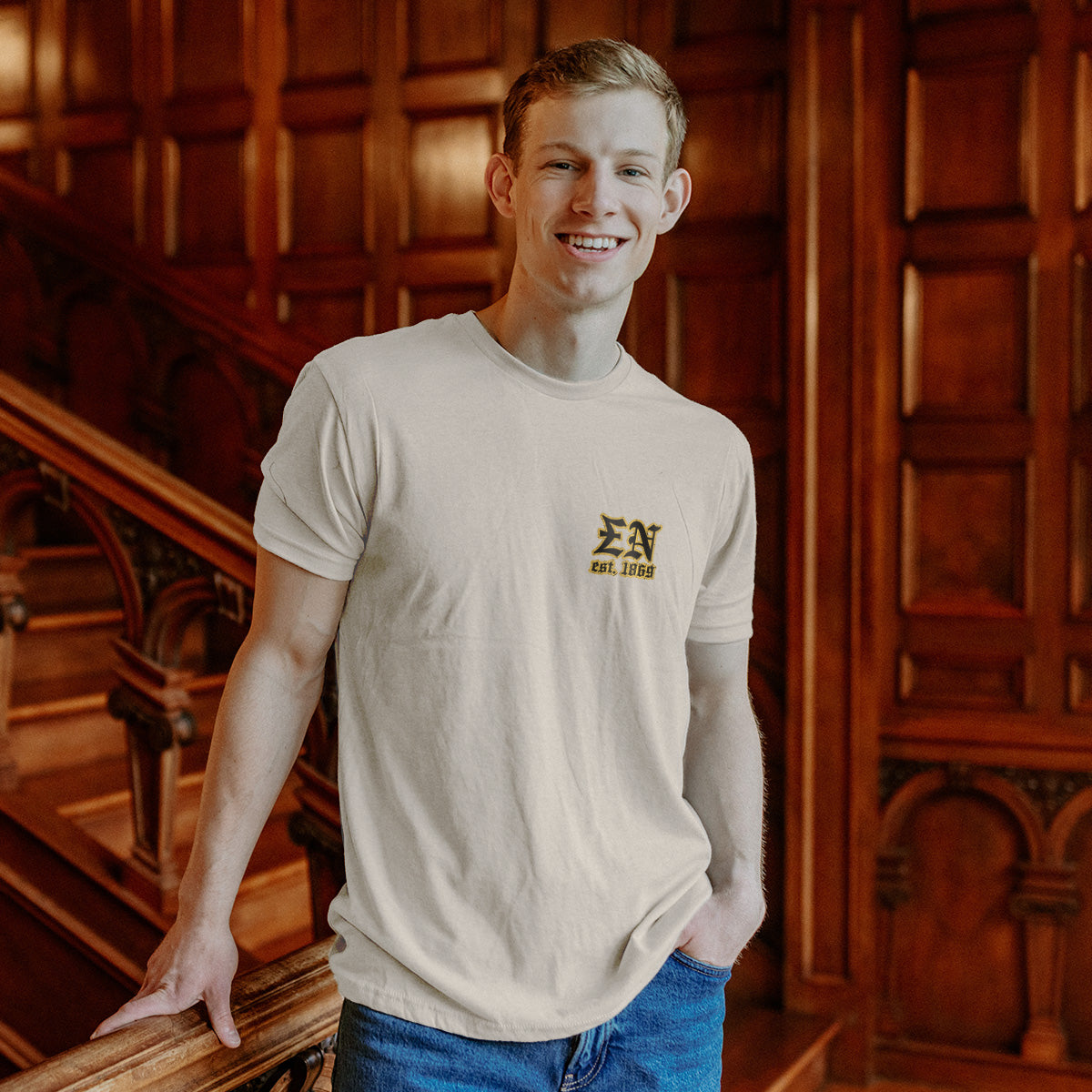 Model wearing the Limited Edition Sigma Nu Sword and Serpent Tee, showing the front pocket design with the Sigma Nu letters and Òest. 1869Ó in gold, in front of ornate wooden paneling.