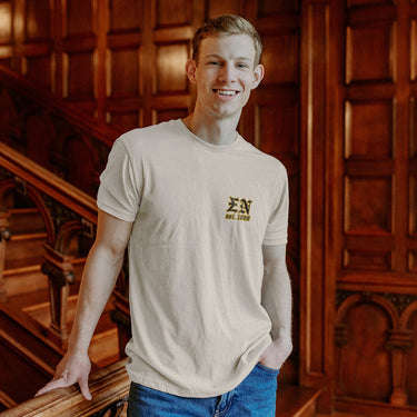 Model wearing the Limited Edition Sigma Nu Sword and Serpent Tee, showing the front pocket design with the Sigma Nu letters and Γest. 1869Γ in gold, in front of ornate wooden paneling.