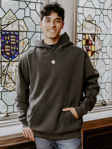 College-aged man wearing the Sigma Nu embroidered black hoodie, standing in front of stained glass windows with fraternity-style crest designs.