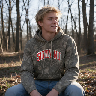 College-aged male wearing the fraternity Deadwood Camo Hoodie with red and white Sigma Nu lettering, seated outdoors in a wooded setting.