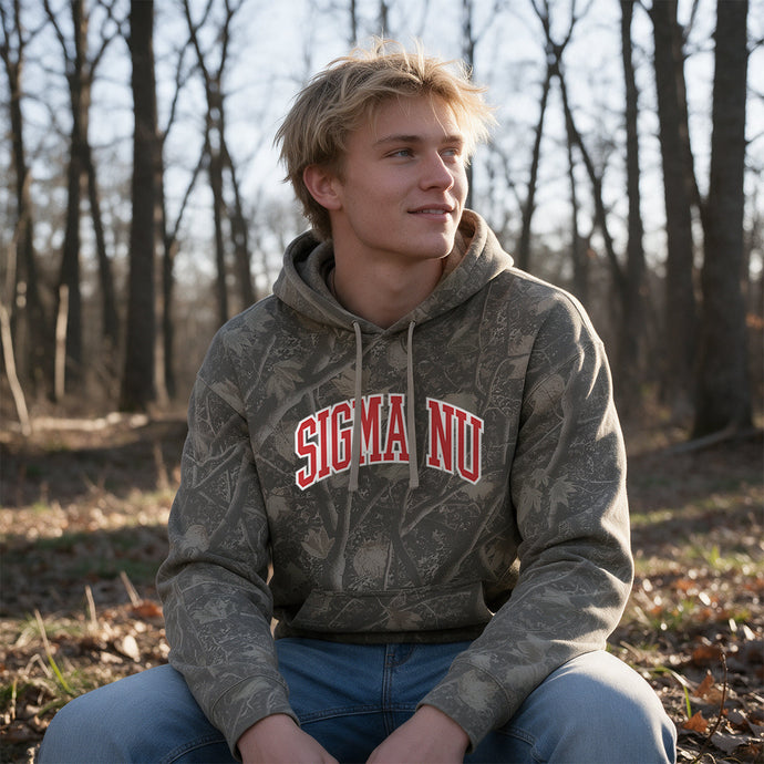 College-aged male wearing the fraternity Deadwood Camo Hoodie with red and white Sigma Nu lettering, seated outdoors in a wooded setting.