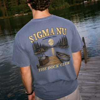 Young man wearing a blue graphic tee standing on a lakeside dock, showing the back design
