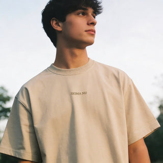 Man wearing a beige crewneck t-shirt with subtle embroidered text on the chest, photographed outdoors.