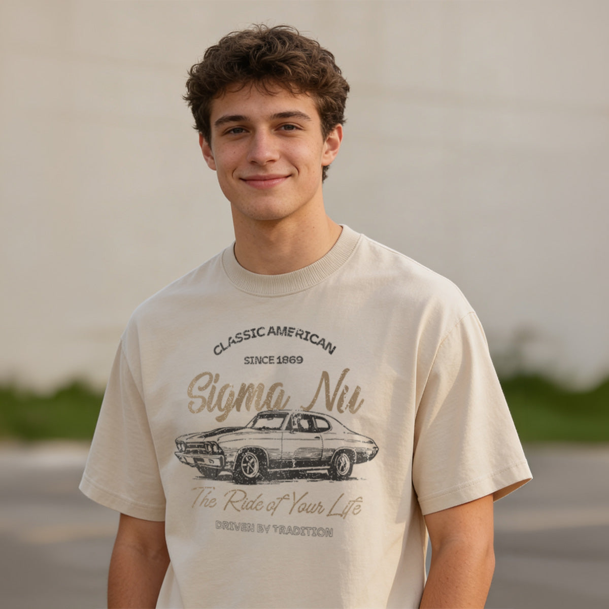 Young man wearing a vintage-style beige graphic T-shirt featuring a retro muscle car illustration and cursive text, standing outdoors with a neutral blurred background.